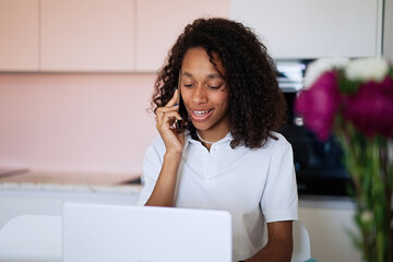 Beautiful young woman sitting at kitchen counter, drinking coffee and chatting on a mobile phone while relaxing at home in the morning. Working on a computer in an office at home