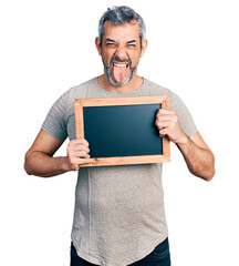 Middle age hispanic man with grey hair holding blackboard sticking tongue out happy with funny...