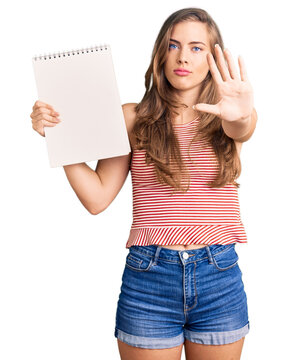 Beautiful caucasian young woman holding notebook with open hand doing stop sign with serious and confident expression, defense gesture