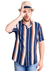Young handsome caucasian man wearing summer hat doing ok gesture shocked with surprised face, eye looking through fingers. unbelieving expression.