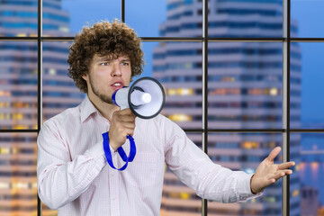 Handsome young man with curly hair in white shirt speaks in megaphone. Indoor window with night...