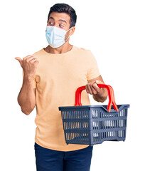 Young hispanic man wearing shopping basket and medical mask pointing thumb up to the side smiling...
