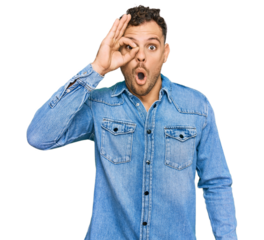 Young hispanic man wearing casual denim jacket doing ok gesture shocked with surprised face, eye looking through fingers. unbelieving expression.