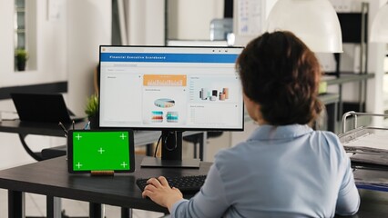 Businesswoman works at desk with greenscreen presented on modern tablet, looking at isolated copyspace template while she analyzes revenue data on pc. Worker uses chromakey display. Handheld shot.
