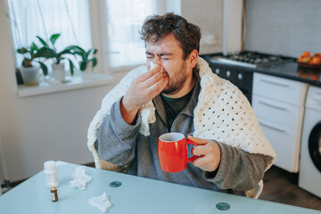 Flu Symptom Management Young Man with Beard in Duvet, Shivering and Wiping Nose at Home in Kitchen