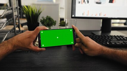 POV of worker holds phone with greenscreen display at desk, working with isolated mockup template...