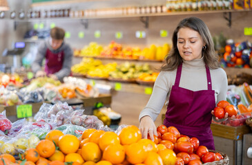 Smiling young saleswoman taking tomatoes from food stall in large grocery store