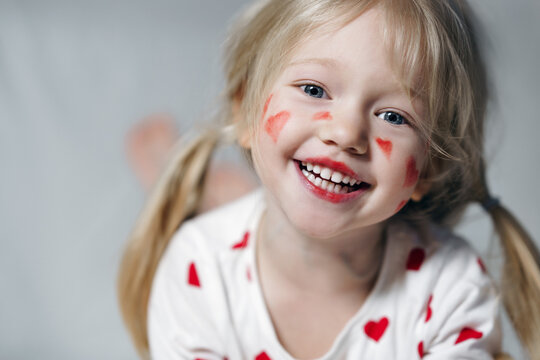 Close up Portrait of positive little sincere adorable kid girl with painted red hearts on face in white pajama with printed red hearts looking at camera on white background. Valentine's concept 
