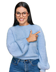 Young hispanic woman wearing casual clothes and glasses cheerful with a smile of face pointing with...