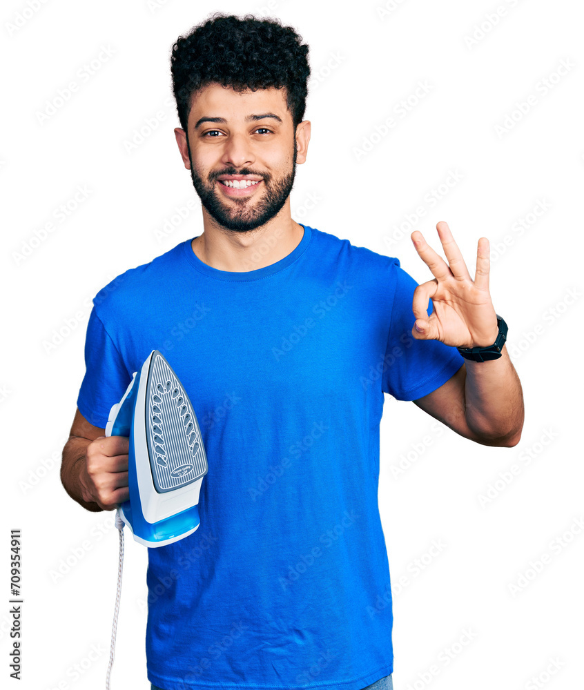 Canvas Prints Young arab man with beard holding electric steam iron doing ok sign with fingers, smiling friendly gesturing excellent symbol