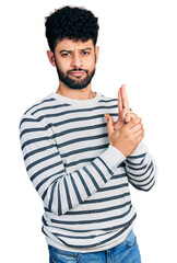 Fototapeta premium Young arab man with beard wearing casual striped sweater holding symbolic gun with hand gesture, playing killing shooting weapons, angry face