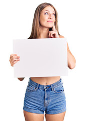 Fototapeta premium Young beautiful blonde woman holding blank empty banner serious face thinking about question with hand on chin, thoughtful about confusing idea