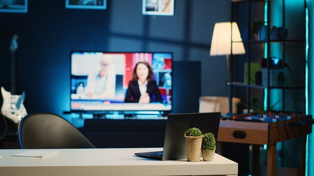 Panning shot of work desk with laptop and mini house plants in dimly lit empty home studio interior. Apartment illuminated by RGB lights with TV news broadcast running in background