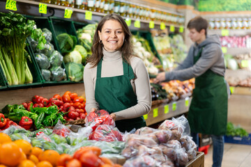 Smiling young saleswoman placing red peppers in polybag on counter in large grocery store