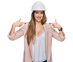 Young blonde woman wearing architect hardhat looking confident with smile on face, pointing oneself with fingers proud and happy.