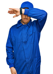 Bald man with beard wearing builder jumpsuit uniform covering eyes with arm, looking serious and sad. sightless, hiding and rejection concept