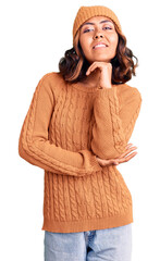 Young beautiful mixed race woman wearing wool sweater and winter hat with hand on chin thinking about question, pensive expression. smiling and thoughtful face. doubt concept.