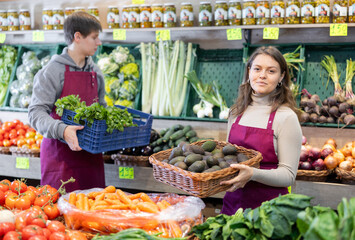 Positive young female seller holding a basket with avocados standing by counter in fruit and...