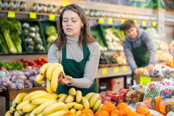 Young woman seller in uniform lays out bananas on counter in grocery store..