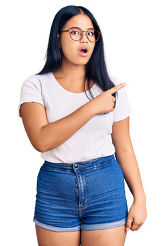 Young beautiful asian girl wearing casual clothes and glasses surprised pointing with finger to the side, open mouth amazed expression.