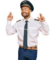 Handsome man with beard wearing airplane pilot uniform gesturing finger crossed smiling with hope and eyes closed. luck and superstitious concept.