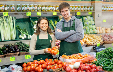 Positive smiling guy and girl sellers offering fresh vegetables in vegetable store
