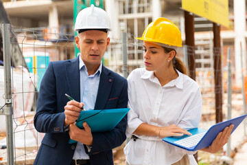 Two young successful architects, controlling the progress of work on the construction site, discuss the construction plan, ..holding a laptop and a folder with drawings in their hands, making