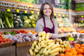 Happy young shop assistant setting bunch of bananas on a counter in big greengrocery