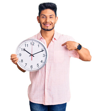 Handsome latin american young man holding big clock pointing finger to one self smiling happy and proud