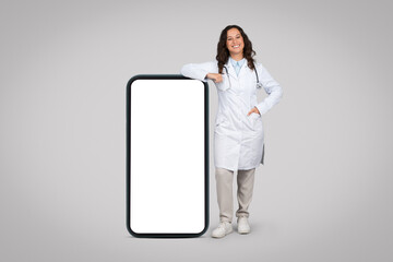 Cheerful young female doctor leaning on giant smartphone with white blank screen standing over grey...