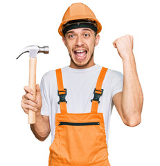 Hispanic young man wearing hardhat holding hammer screaming proud, celebrating victory and success very excited with raised arms