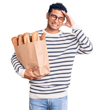 Hispanic handsome young man holding paper bag with bread stressed and frustrated with hand on head, surprised and angry face