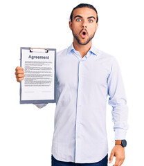 Young handsome man holding clipboard with agreement document scared and amazed with open mouth for surprise, disbelief face