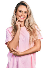 Young blonde woman wearing casual pink t shirt smiling looking confident at the camera with crossed arms and hand on chin. thinking positive.