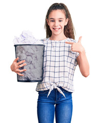 Cute hispanic child girl holding paper bin full of crumpled papers pointing finger to one self smiling happy and proud