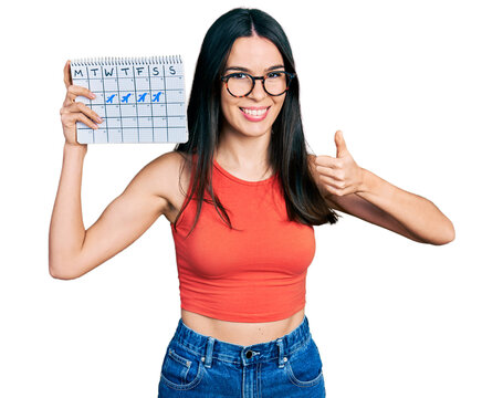 Young hispanic woman holding travel calendar smiling happy and positive, thumb up doing excellent and approval sign