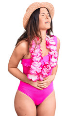 Young beautiful brunette woman wearing swimwear and hawaiian lei with hand on stomach because...