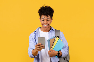 Joyful student using smartphone with notebooks on yellow