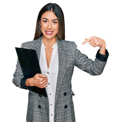 Young hispanic woman wearing business clothes holding clipboard pointing finger to one self smiling...
