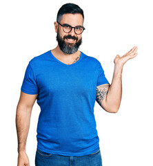 Hispanic man with beard wearing casual t shirt and glasses smiling cheerful presenting and pointing with palm of hand looking at the camera.