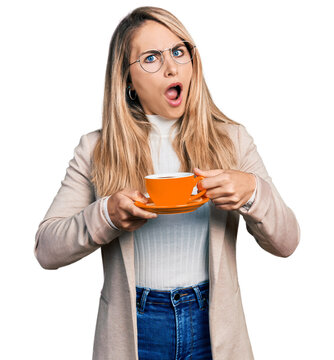 Young blonde woman wearing business style drinking cup of coffee in shock face, looking skeptical and sarcastic, surprised with open mouth