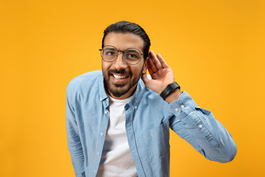 A jovial man with a beard and glasses leans in with a hand to his ear, portraying a listening or hearing action