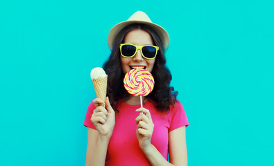 Summer portrait of happy young woman eating lollipop and ice cream wearing sunglasses and straw tourist hat on blue background