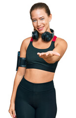 Beautiful blonde woman wearing gym clothes and using headphones smiling cheerful offering palm hand giving assistance and acceptance.