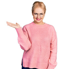 Young blonde woman wearing casual clothes and glasses smiling cheerful presenting and pointing with palm of hand looking at the camera.