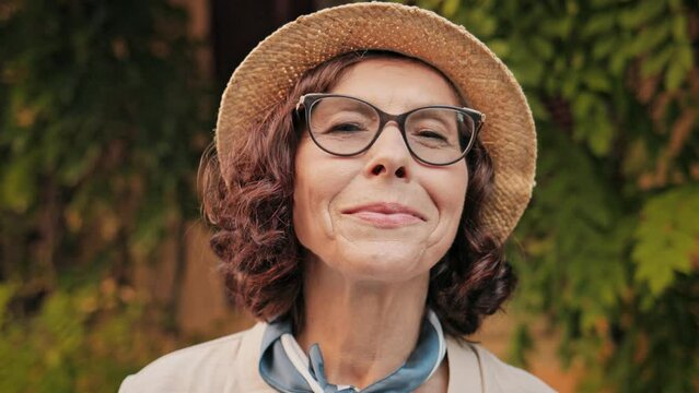 Very close camera view of beautiful Caucasian woman in middle of street looking to side upwards and afterwards to camera. Beginning to smile with joy. Wearing glasses and hat. Greenery in background.
