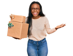 Young black woman holding gifts celebrating victory with happy smile and winner expression with raised hands