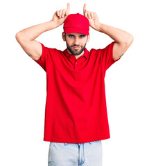 Young handsome man with beard wearing delivery uniform doing funny gesture with finger over head as bull horns