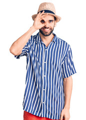 Young handsome man with beard wearing summer hat and striped shirt doing ok gesture with hand smiling, eye looking through fingers with happy face.