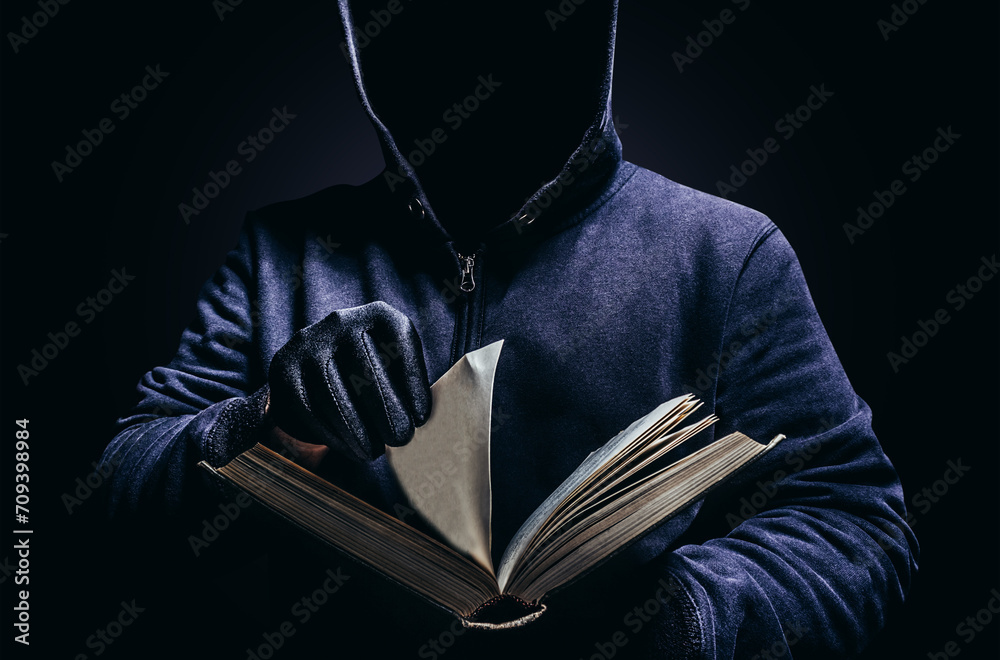 Wall mural Photo of scary shaded hooded man holding book and turning the pages on dark background. Horror book concept.